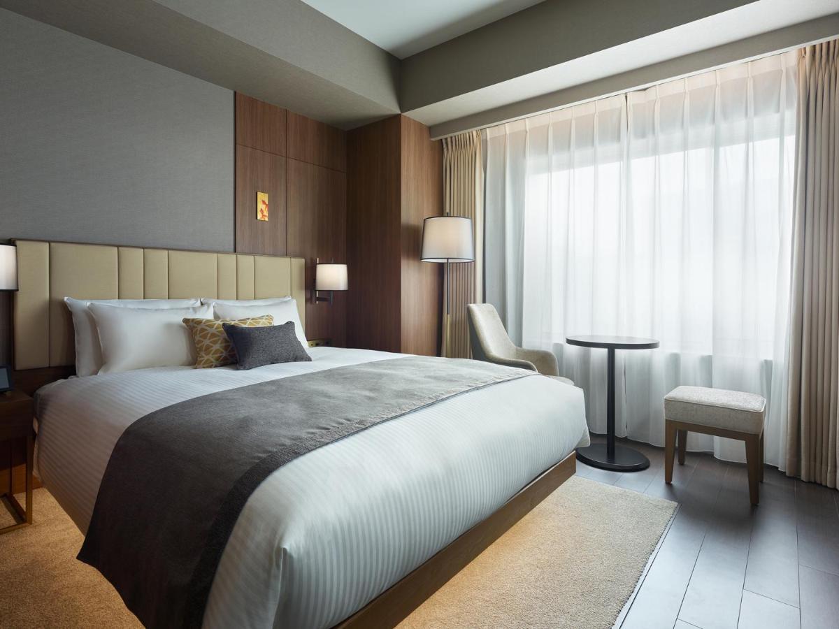 Mitsui Garden Hotel Roppongi Tokyo Premier Dış mekan fotoğraf The photo shows a modern hotel room featuring a comfortable bed with white linens and a decorative throw pillow. The room has a neutral color palette with wooden accents on the walls. There are two bedside lamps and a chair positioned next to a small