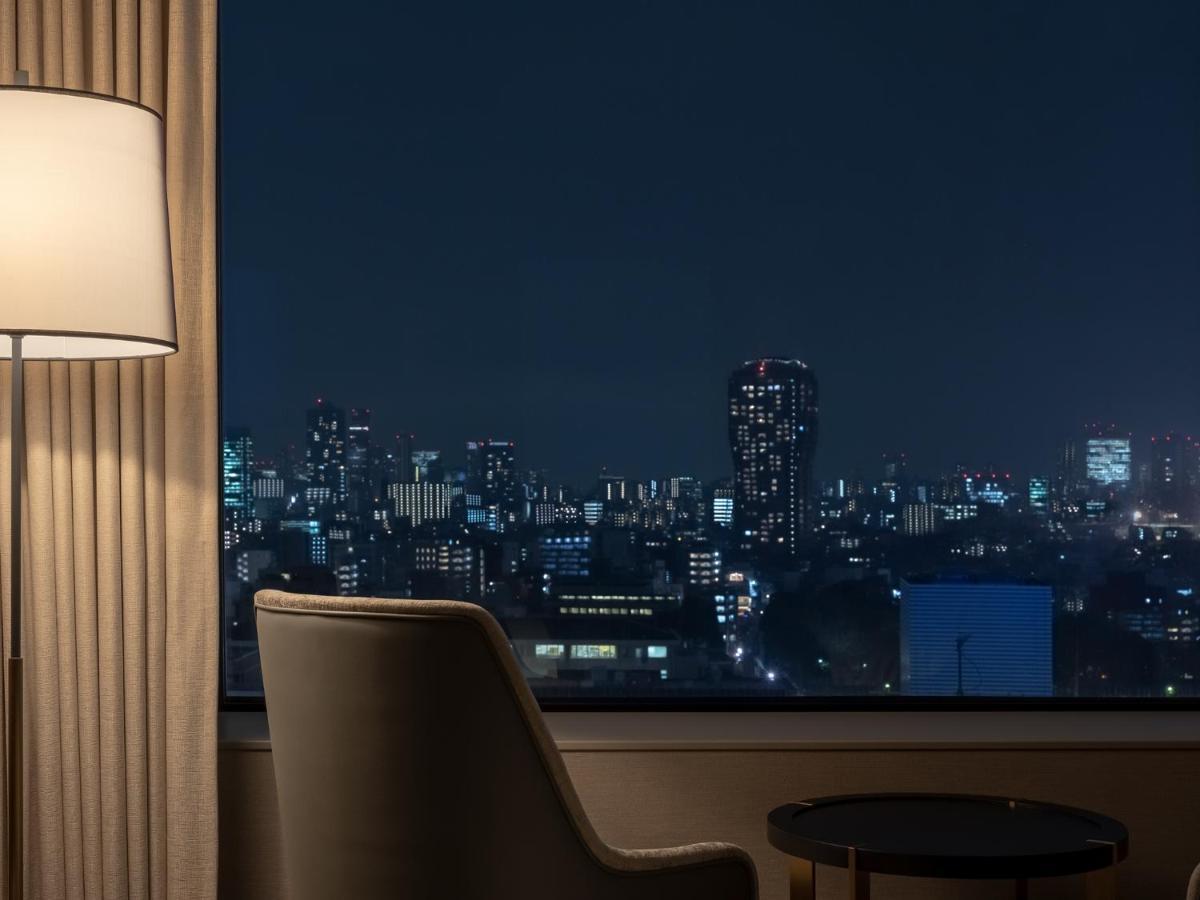 Mitsui Garden Hotel Roppongi Tokyo Premier Dış mekan fotoğraf The photo depicts a cozy hotel room setting at night. In the foreground, there's a stylish armchair next to a small round table. A standing lamp provides soft lighting, creating a warm ambiance. In the background, large windows reveal a city skyline 
