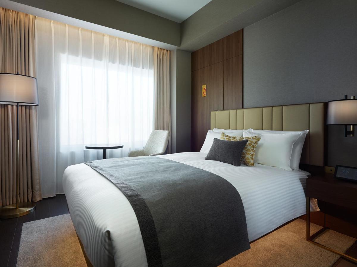 Mitsui Garden Hotel Roppongi Tokyo Premier Dış mekan fotoğraf The photo shows a modern hotel room featuring a neatly made bed with white linens and a decorative gray throw blanket. To the side, there is a small round table and a comfortable chair, creating a cozy seating area. The room is well-lit with natural 