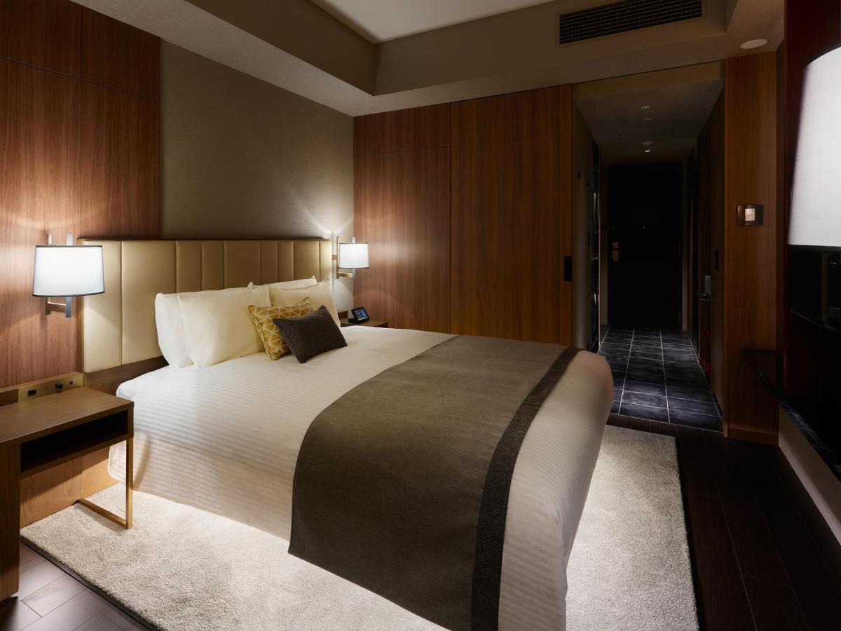Mitsui Garden Hotel Roppongi Tokyo Premier Dış mekan fotoğraf The image shows a modern hotel room with a warm and elegant design. In the center, there is a neatly made bed with white linens and a dark gray throw blanket. Flanking the bed are two bedside lamps providing soft lighting. The walls have a rich wood 