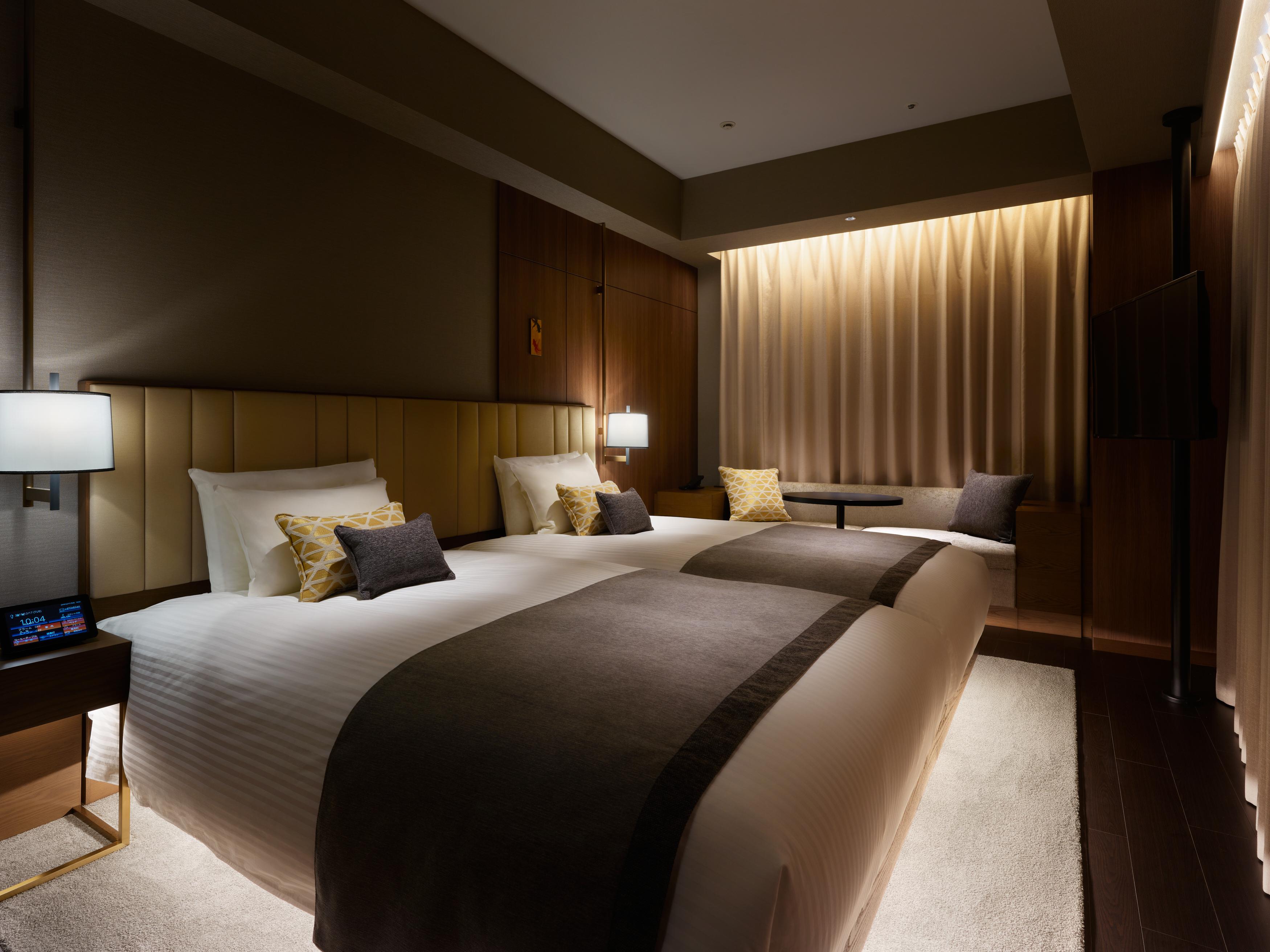 Mitsui Garden Hotel Roppongi Tokyo Premier Dış mekan fotoğraf The photo shows a modern hotel room featuring two beds with plush white linens and decorative pillows. The room has a warm and inviting ambiance, highlighted by soft lighting from lamps and the backlit curtains. There is also a small table with chair