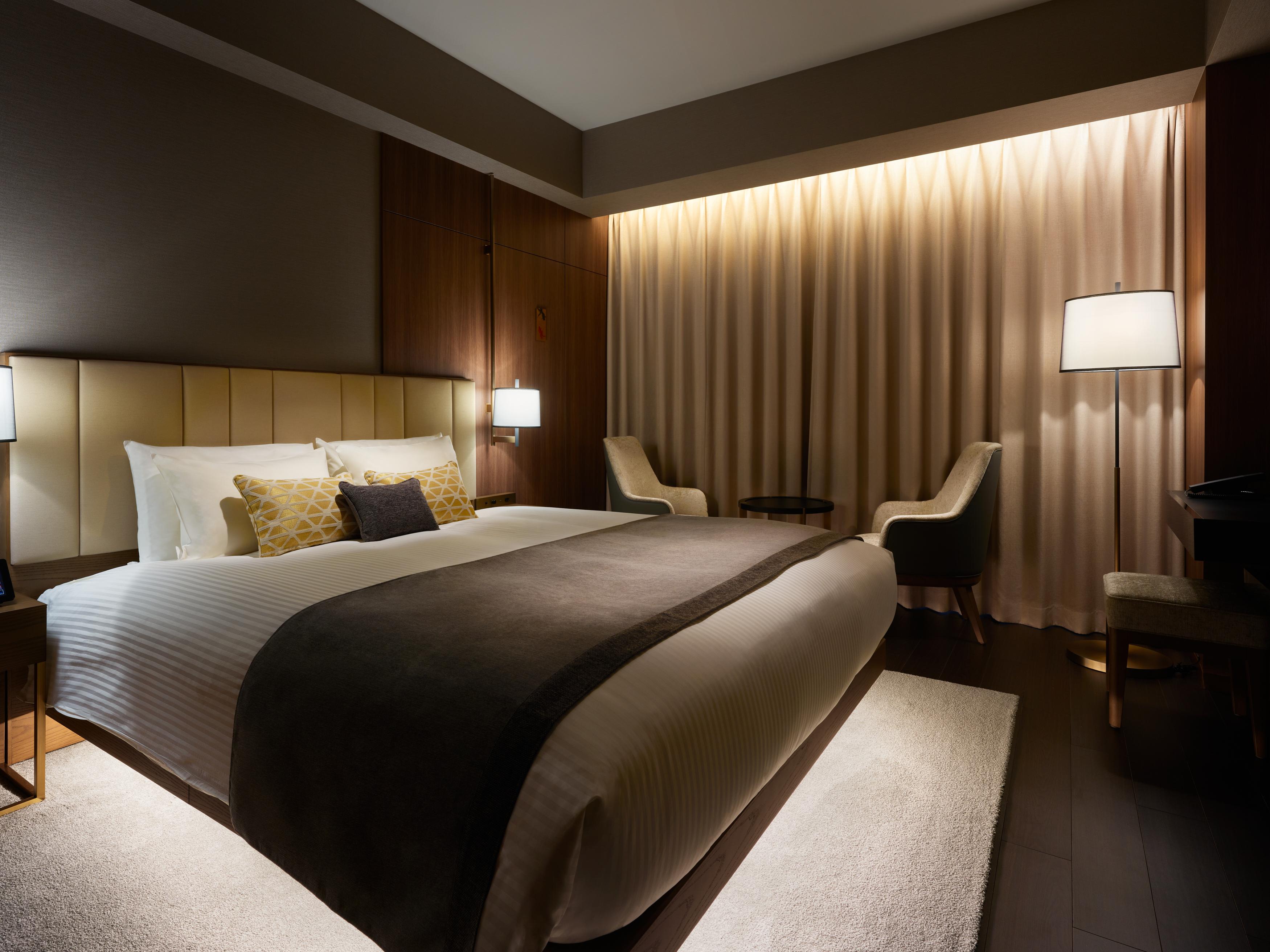 Mitsui Garden Hotel Roppongi Tokyo Premier Dış mekan fotoğraf The photo depicts a modern hotel bedroom. It features a large bed with white linens and a dark gray throw blanket, accented by decorative pillows. The room has a warm ambiance created by soft lighting, including two bedside lamps and a lighted area b