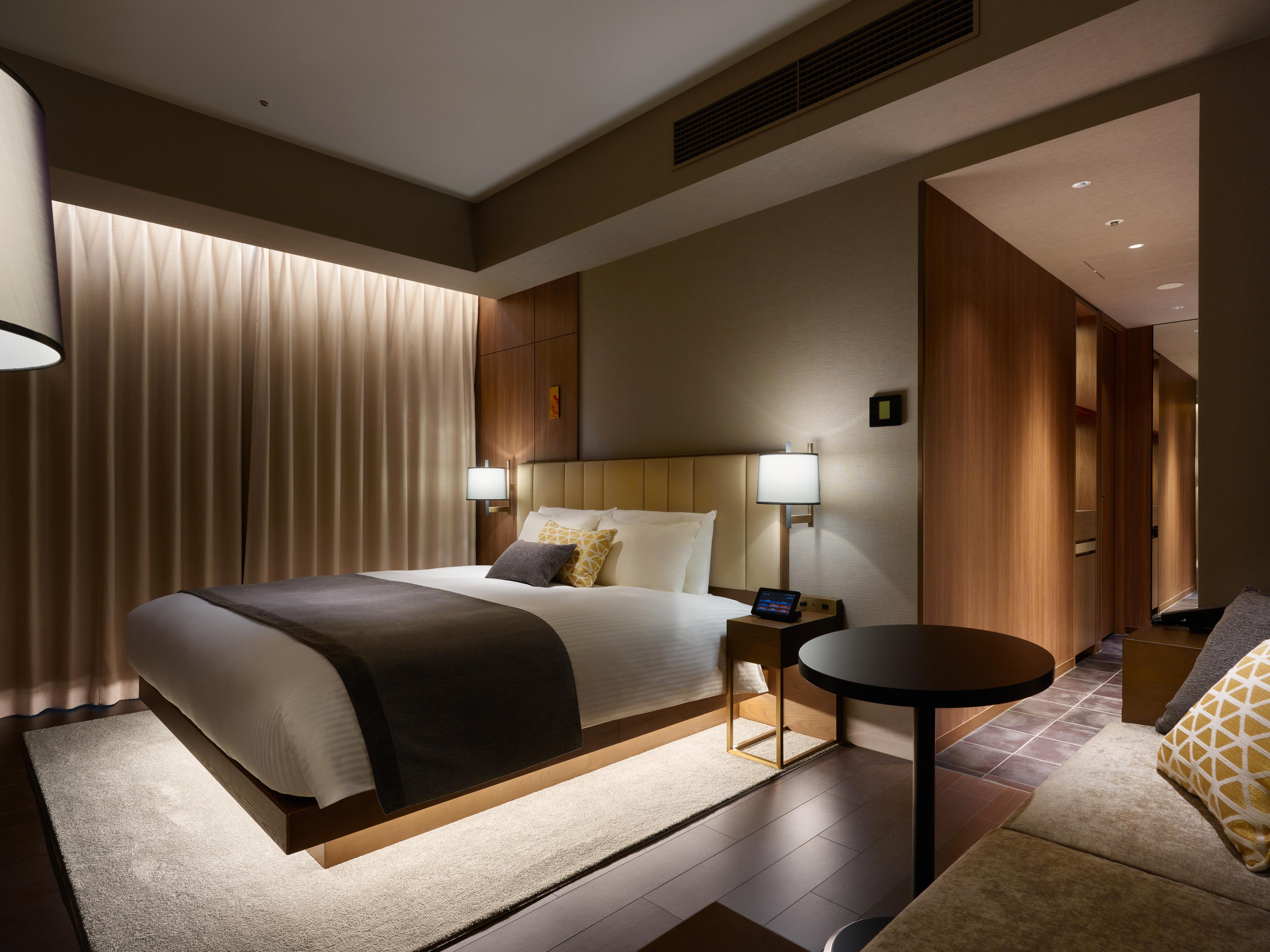 Mitsui Garden Hotel Roppongi Tokyo Premier Dış mekan fotoğraf The photo shows a modern hotel room featuring a king-sized bed with neutral-colored bedding and accent pillows. There are two bedside lamps on either side of the bed, providing a warm glow. The walls have a wood finish, and there's a soft area rug on