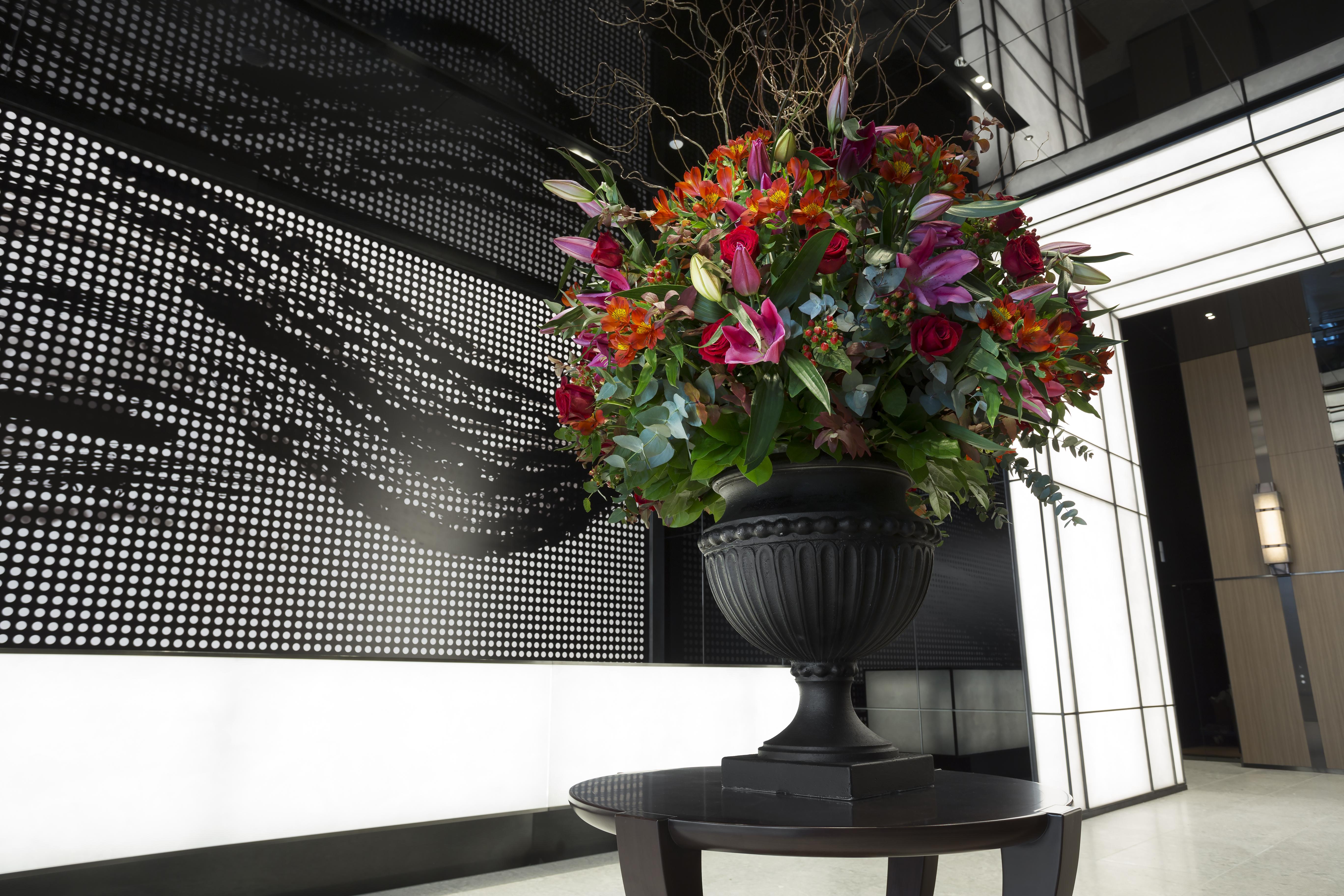 Mitsui Garden Hotel Roppongi Tokyo Premier Dış mekan fotoğraf The photo features a large, vibrant floral arrangement in an ornate black urn. The arrangement is composed of various colorful flowers and greenery, showcasing shades of red, pink, orange, and green. This floral display is positioned on a small table