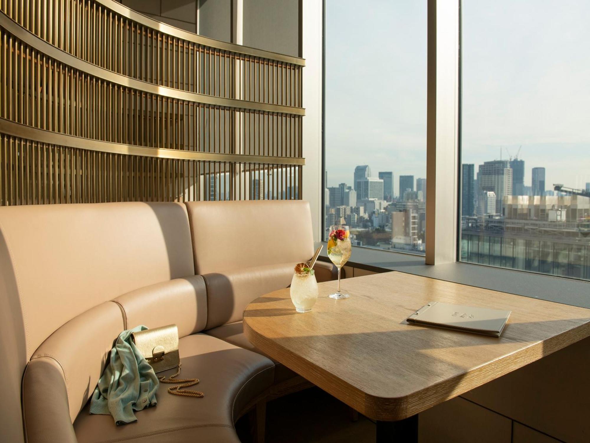 Mitsui Garden Hotel Roppongi Tokyo Premier Dış mekan fotoğraf The photo depicts a cozy, modern seating area with a curved booth that features soft, pale upholstery. A wooden table is positioned in front of the booth, and on the table, there are two elegant drinks garnished with fruit. The setting has large wind