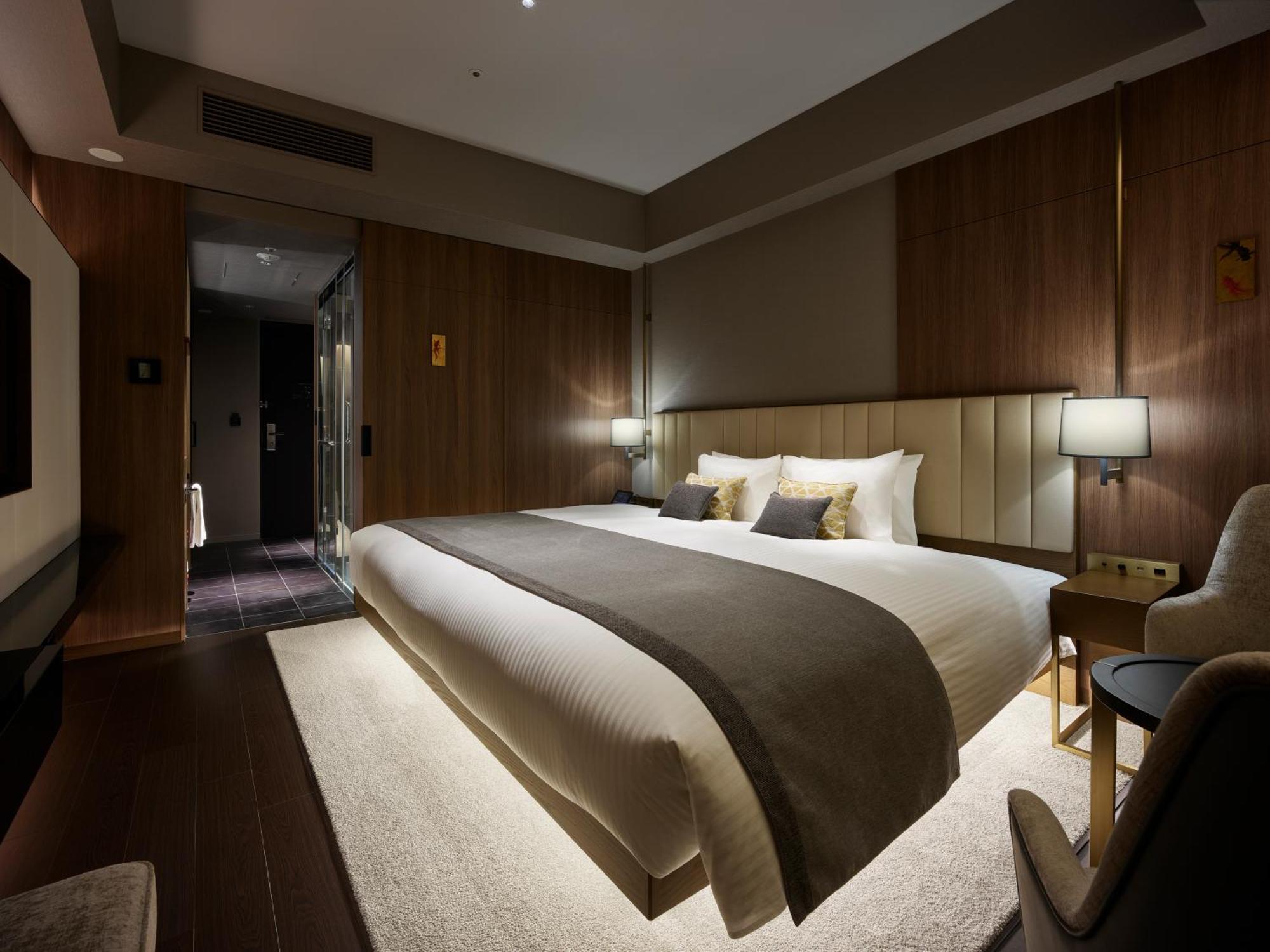 Mitsui Garden Hotel Roppongi Tokyo Premier Dış mekan fotoğraf The photo shows a modern hotel room featuring a large bed with a neutral-colored duvet and a dark throw blanket. The headboard is upholstered, and there are two bedside lamps on either side of the bed. The walls have a warm wood paneling, contributin