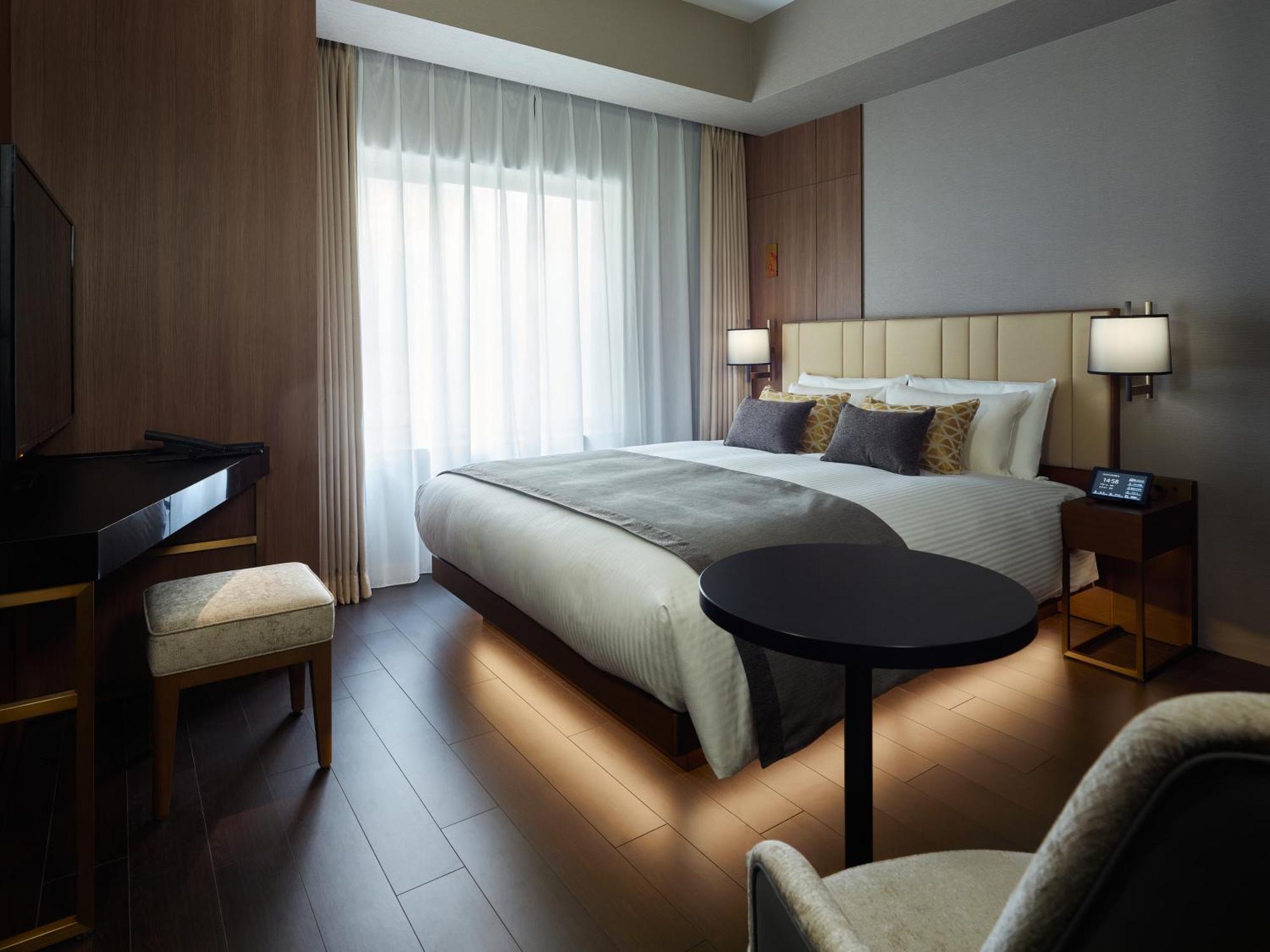 Mitsui Garden Hotel Roppongi Tokyo Premier Dış mekan fotoğraf The photo shows a modern hotel room featuring a large bed with decorative pillows. There are large windows with sheer curtains allowing natural light to filter in. To one side, there is a sleek desk with a chair and a small round table in the center 