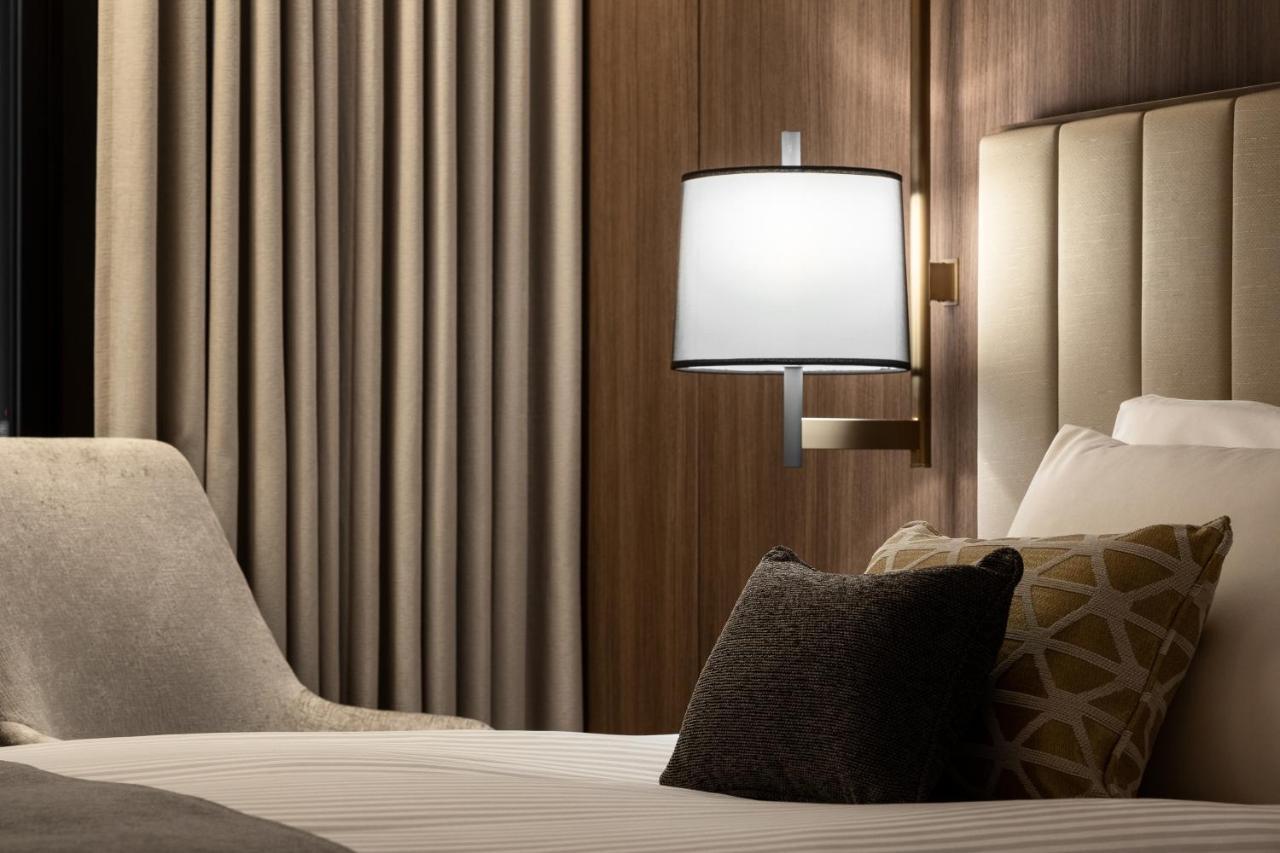 Mitsui Garden Hotel Roppongi Tokyo Premier Dış mekan fotoğraf The photo shows a cozy hotel bedroom setting. There is a neatly made bed with white linens and a few decorative pillows, including a dark one and another with a geometric pattern. To the side, there is a soft armchair and a modern wall lamp mounted a